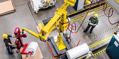 Overhead image of a worker operating a machine while another stands in an area marked off with black-and-yellow stripe tape

