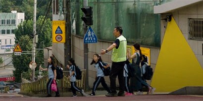 산이 많은 도심 지역에서 어린이들이 형광 안전조끼를 입은 어른의 지시에 따라 길을 건넙니다.
