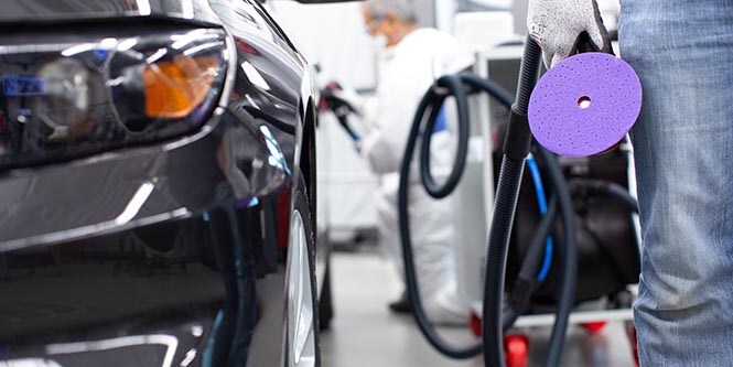 Photograph showing a close-up of a black car's front side with a person holding a purple polishing pad and a hose.
