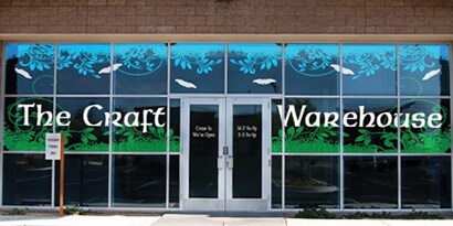 Exterior of a business with glass walls and doors with a graphic spelling out The Craft Warehouse with a blue and green colored film background using 3M graphic colored films.
