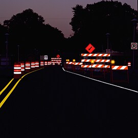 Temporary traffic control devices in a nighttime road work zone are illuminated by headlights.