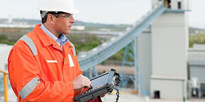  Image of an engineer in safety gear entering information into a hand held computer while looking out over an industrial plant.
